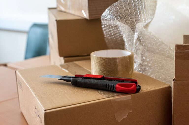 Bastelmesser und durchsichtiges Klebeband auf dem Verpackungskarton auf dem Schreibtisch im Versandbüro